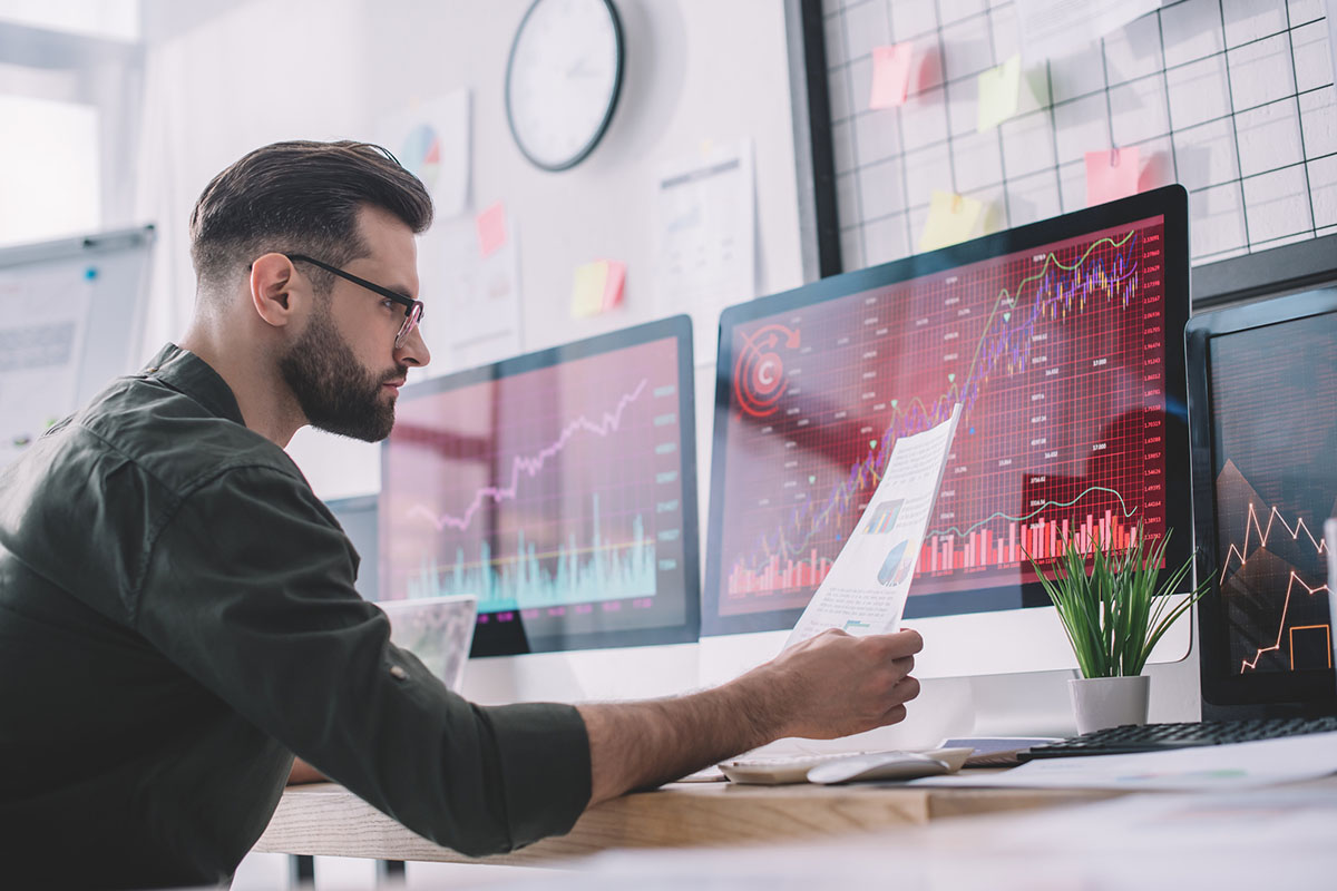 A man is studying charts and graphs on a dual-screen monitor desktop setup.