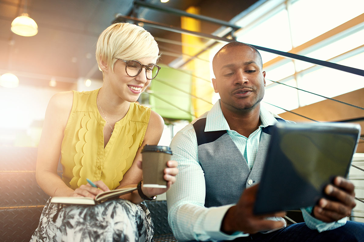 A young woman with short blonde hair and glasses smiles while holding a coffee cup and writing in a notebook. Beside her, a man in a gray vest looks at a tablet, engaged in conversation. They are seated on stairs in a well-lit modern space.