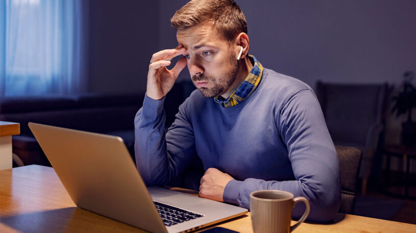 A man with short hair sits at a desk, looking intently at a laptop screen while resting his forehead on his hand. He wears a blue sweater with a collared shirt that has a checkered pattern. A beige mug and a smartphone are on the table in front of him, and the room is dimly lit.