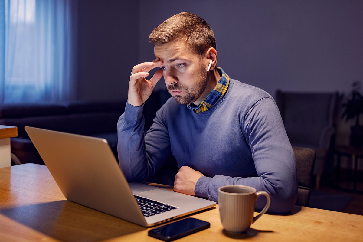 A man with short hair sits at a desk, looking intently at a laptop screen while resting his forehead on his hand. He wears a blue sweater with a collared shirt that has a checkered pattern. A beige mug and a smartphone are on the table in front of him, and the room is dimly lit.