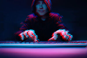 A person in a dark hoodie is typing on a keyboard, with a blurred effect giving a sense of movement. The scene is illuminated with blue and red tones, enhancing the mysterious atmosphere.