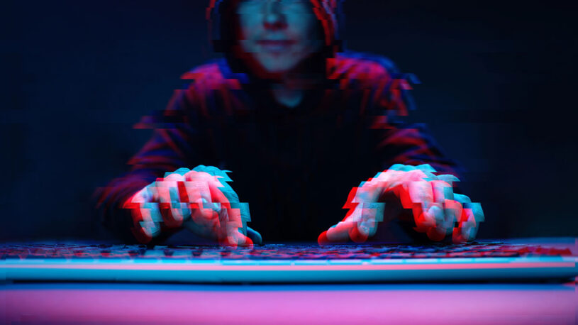A person in a dark hoodie is typing on a keyboard, with a blurred effect giving a sense of movement. The scene is illuminated with blue and red tones, enhancing the mysterious atmosphere.
