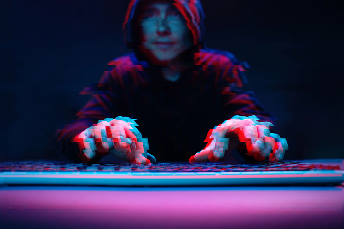 A person in a dark hoodie is typing on a keyboard, with a blurred effect giving a sense of movement. The scene is illuminated with blue and red tones, enhancing the mysterious atmosphere.