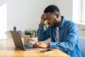 A man with short hair sits at a desk, looking intently at a laptop screen while resting his forehead on his hand. He wears a blue sweater with a collared shirt that has a checkered pattern. A beige mug and a smartphone are on the table in front of him.