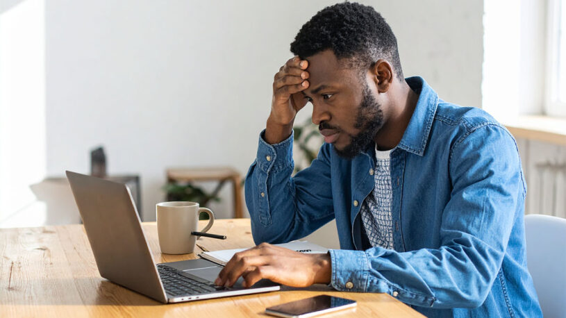A man with short hair sits at a desk, looking intently at a laptop screen while resting his forehead on his hand. He wears a blue sweater with a collared shirt that has a checkered pattern. A beige mug and a smartphone are on the table in front of him.