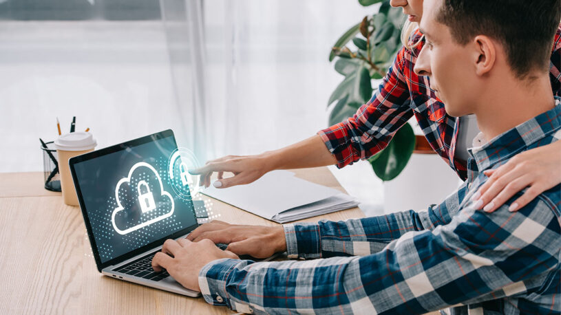 A man and a woman are interacting with a laptop on a wooden table, with digital cloud and lock icons displayed on the screen, symbolizing cybersecurity. The woman is pointing at the screen while placing her hand on the man's shoulder. A coffee cup and stationery are visible in the background.