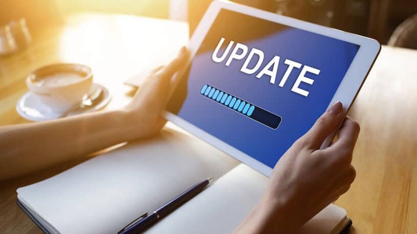 A person holding a tablet displaying a blue screen with the word "UPDATE" and a loading progress bar, with a notebook and a cup of coffee in the background.