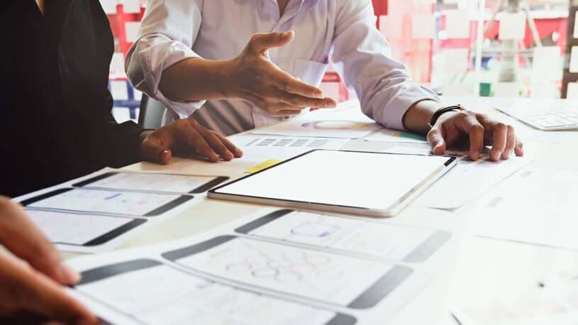 Two individuals are collaborating at a table covered with design mockups for mobile applications, including wireframes and graphs, alongside a tablet displaying a blank screen. One person is gesturing while discussing ideas.
