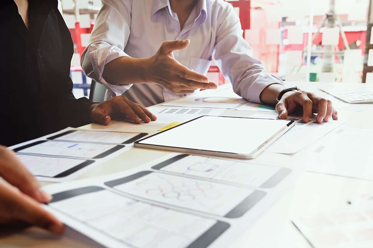 Two individuals are collaborating at a table covered with design mockups for mobile applications, including wireframes and graphs, alongside a tablet displaying a blank screen. One person is gesturing while discussing ideas.