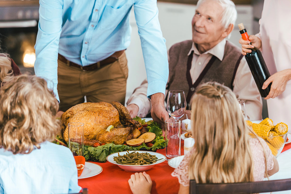 A festive dining table with a roasted turkey at the center, surrounded by various side dishes. A man in a blue shirt serves the turkey while an elderly man in a sweater looks on. Children with curly hair and a girl with long hair sit at the table, watching the scene. A woman pours wine from a bottle.