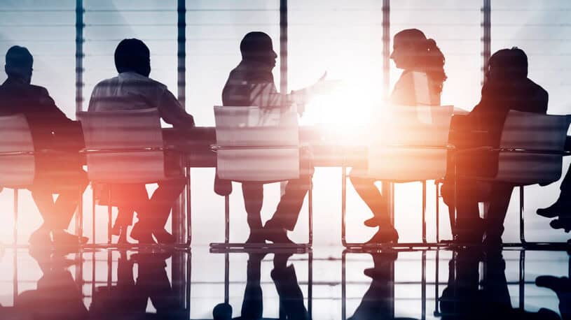 Silhouettes of six individuals seated around a conference table, with sunlight streaming through large windows, creating a dramatic effect on the scene.