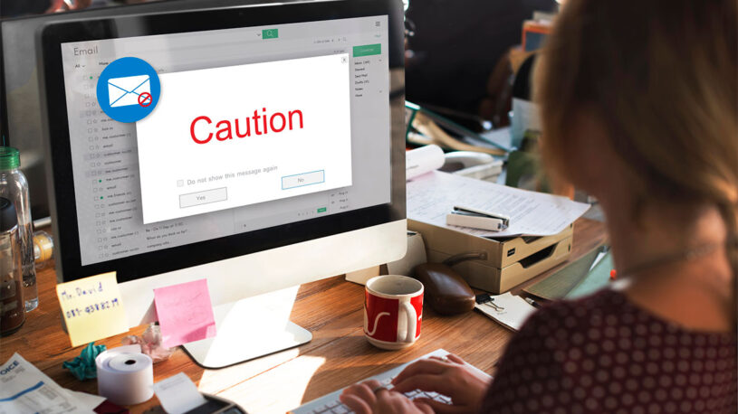 A person is typing on a keyboard in front of a computer displaying an email warning message that says "Caution" in red. The desk is cluttered with paperwork, a red mug, and various office supplies.