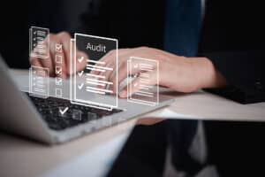 A close-up of hands typing on a laptop with digital audit checklists and documents displayed above the keyboard.