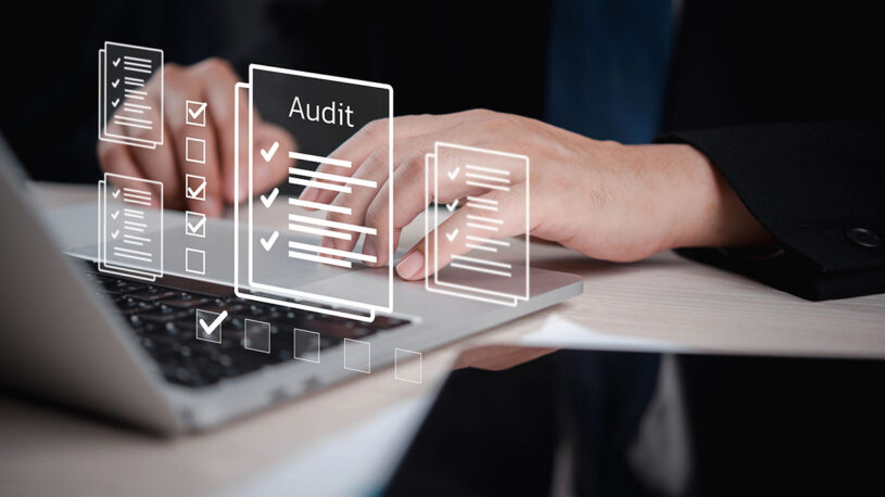 A close-up of hands typing on a laptop with digital audit checklists and documents displayed above the keyboard.
