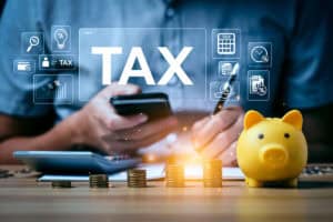 A person holding a smartphone is calculating taxes with a calculator and writing notes, surrounded by stacks of coins and a yellow piggy bank, with graphical tax-related icons displayed above.