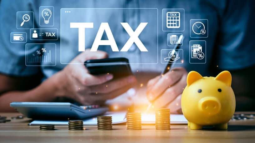 A person holding a smartphone is calculating taxes with a calculator and writing notes, surrounded by stacks of coins and a yellow piggy bank, with graphical tax-related icons displayed above.