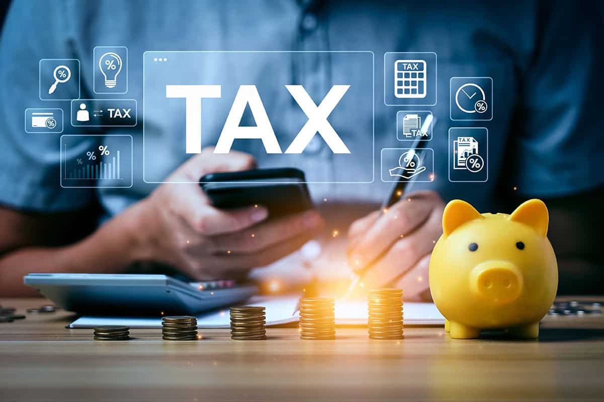 A person holding a smartphone is calculating taxes with a calculator and writing notes, surrounded by stacks of coins and a yellow piggy bank, with graphical tax-related icons displayed above.