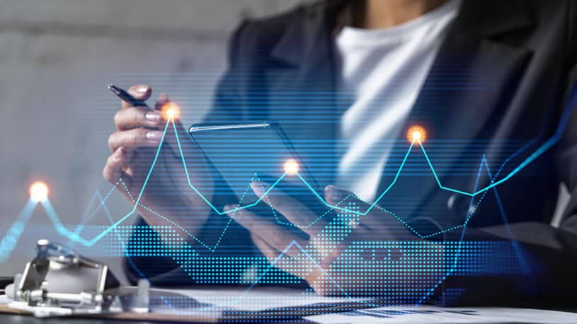 A person in a black blazer holds a smartphone in one hand and a pen in the other, surrounded by digital graphs and data visualizations on a blurred background. Papers and a clipboard are visible on the table.