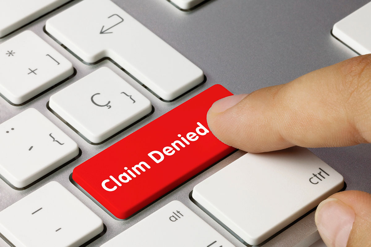 A close-up of a computer keyboard featuring a large red key labelled "Claim Denied," with a finger pressing down on it.