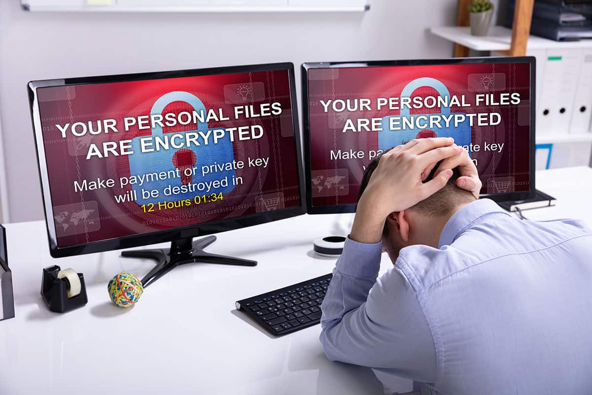 A man sits at a desk with his head down, appearing stressed. Two computer monitors display a warning message: "YOUR PERSONAL FILES ARE ENCRYPTED" and a countdown timer, indicating 12 hours and 1 minute remaining to make a payment or risk losing files. Office supplies are visible on the desk.
